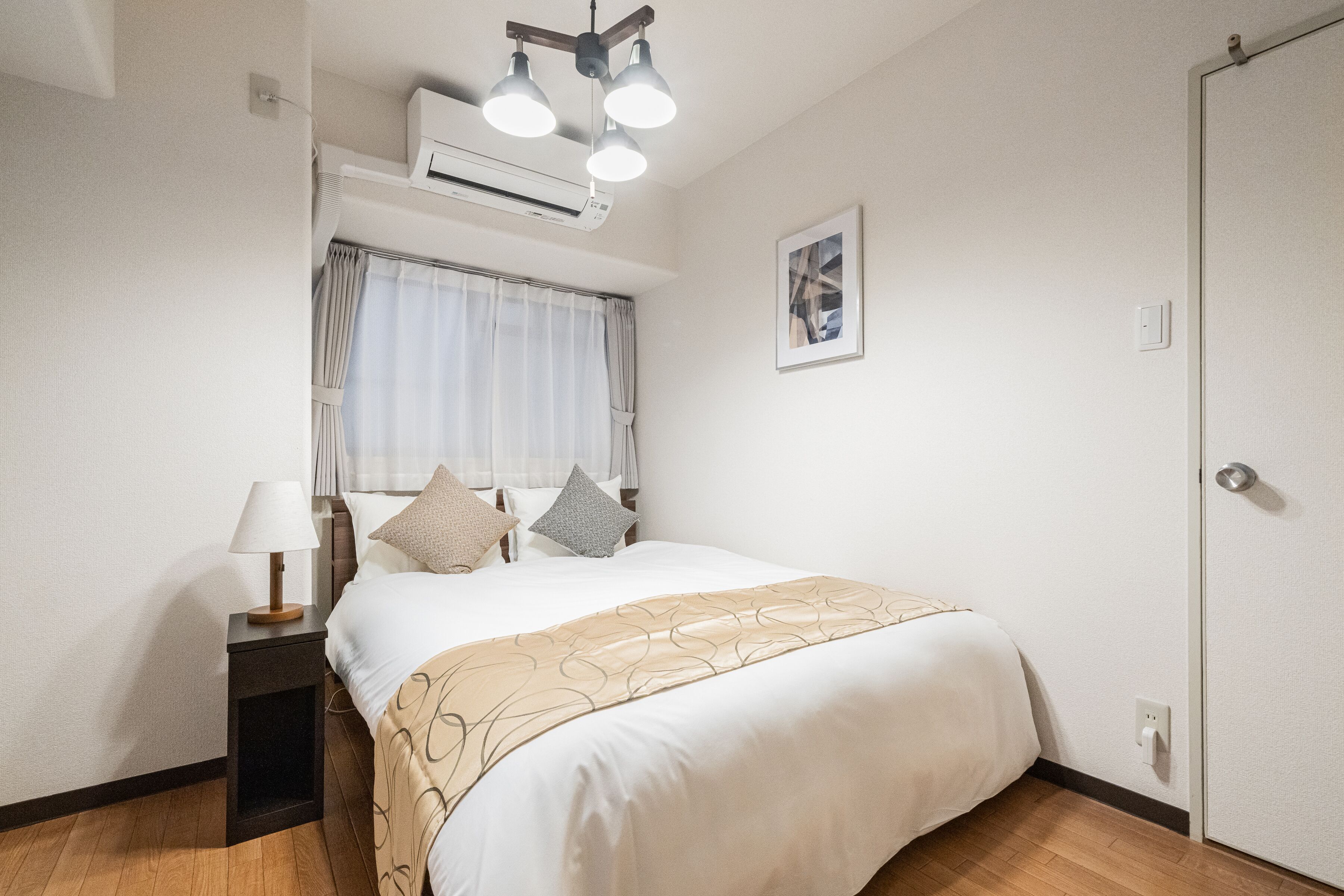 A hotel room with a bed, bedside table, lamp, air conditioning unit, window with curtains, and a door.