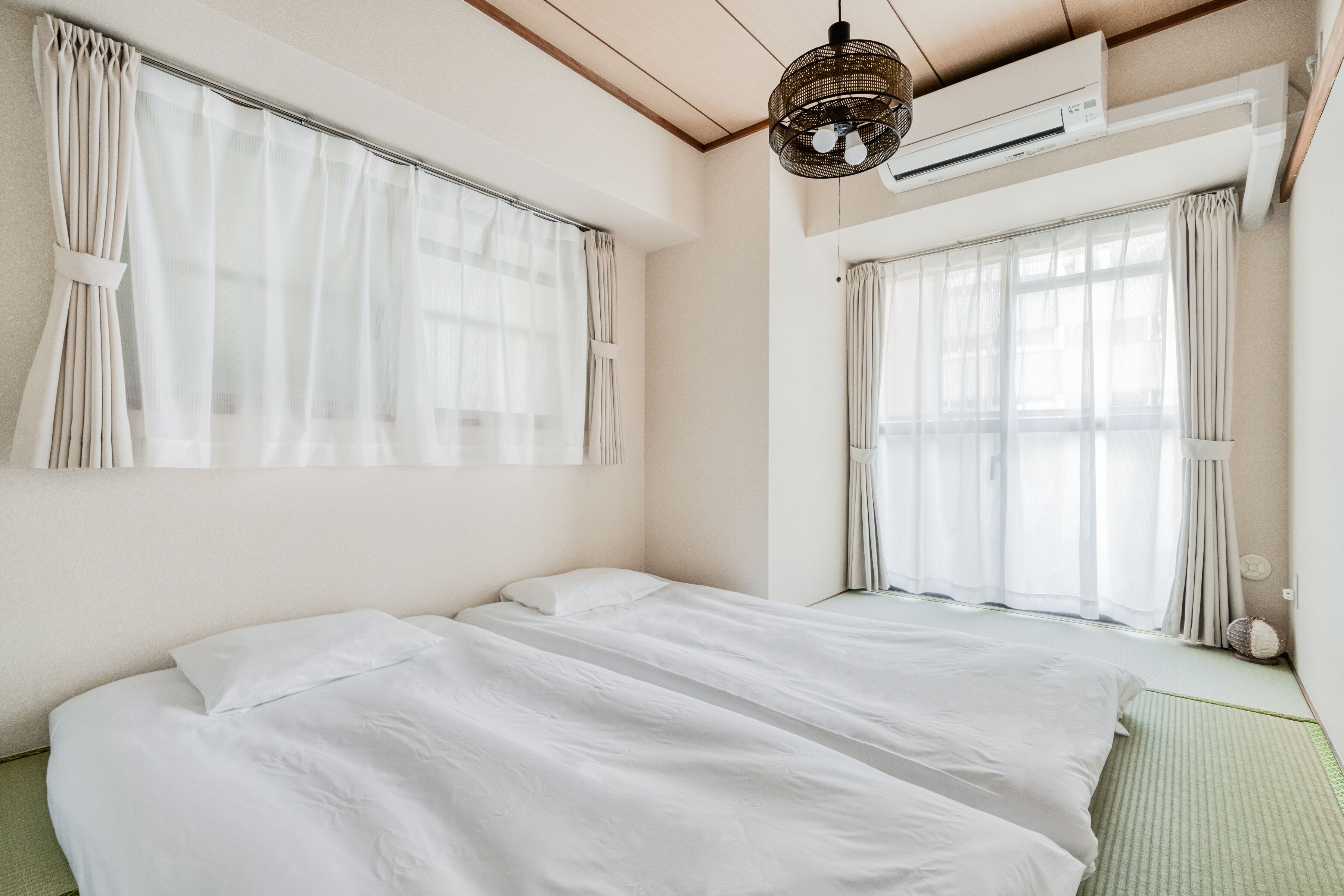 A room with a large bed, two windows with sheer curtains, a ceiling fan, and an air conditioning unit.