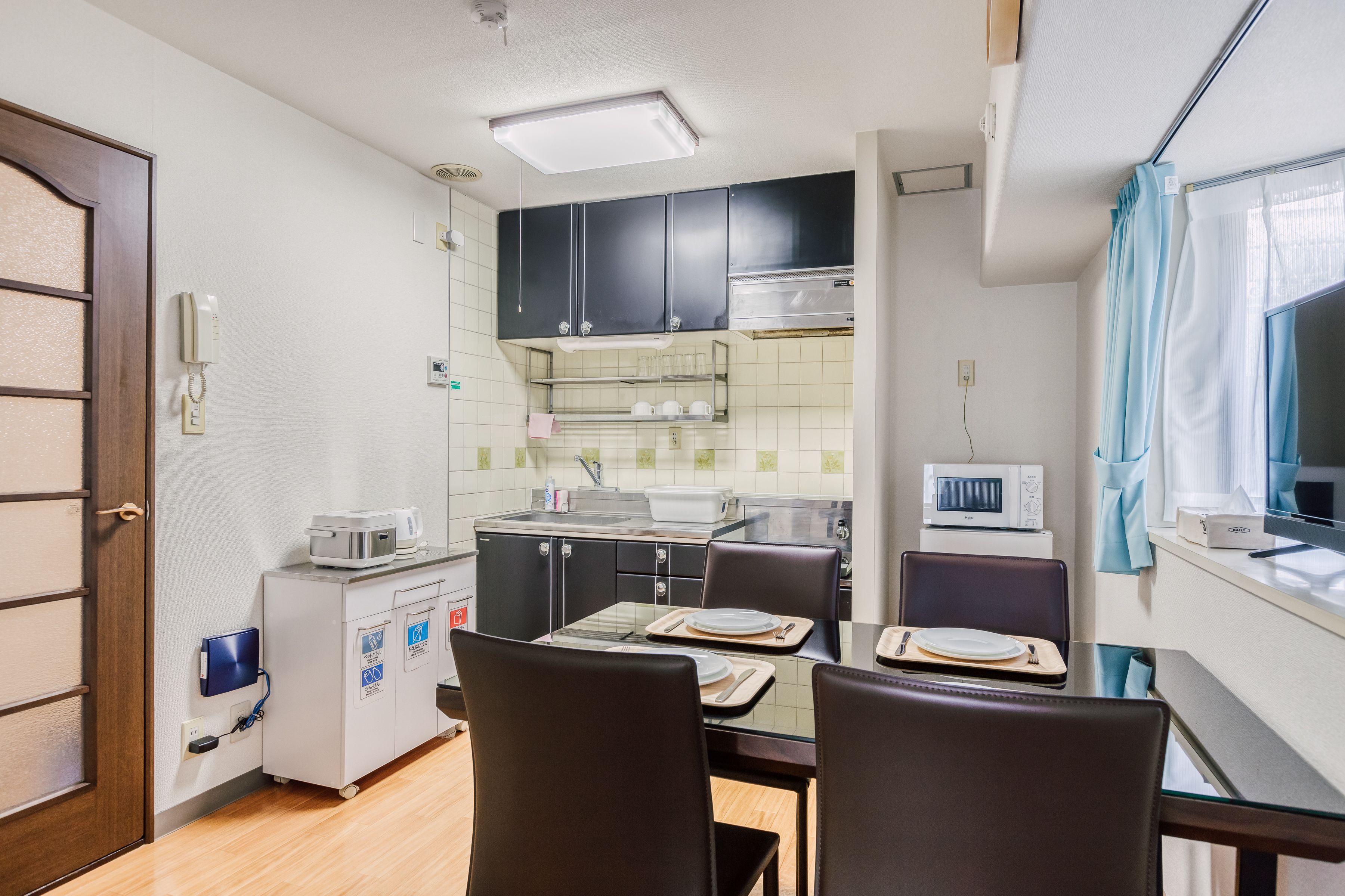 A compact kitchen with a microwave, toaster, and a small refrigerator. There is a dining table with chairs and a window with curtains.