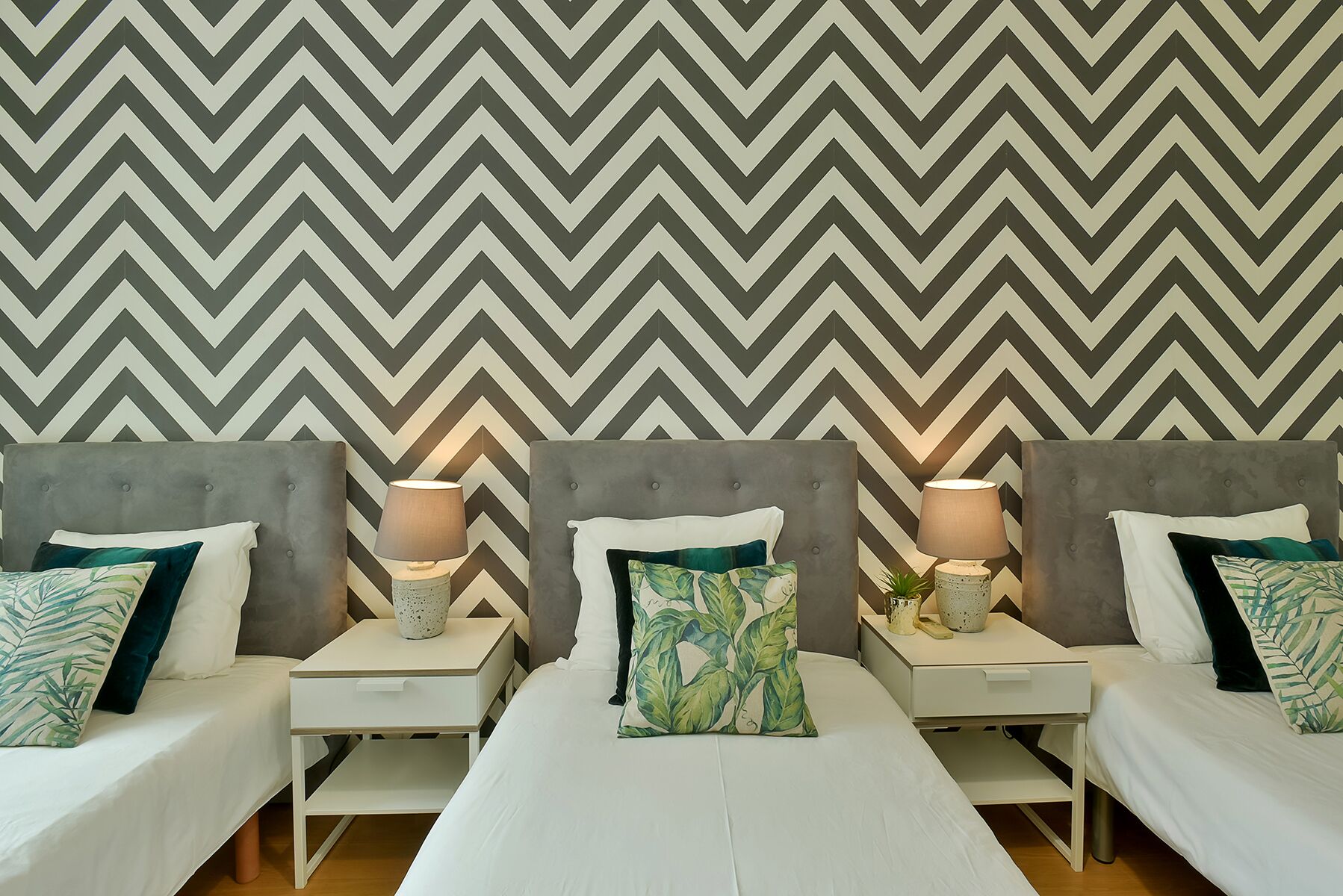 Une chambre d’hôtel avec trois lits, chacun doté d’une tête de lit grise et de linge de lit blanc. On y trouve des tables de chevet avec des lampes et des coussins décoratifs. Le mur est orné d’un grand papier peint à motifs chevrons.
