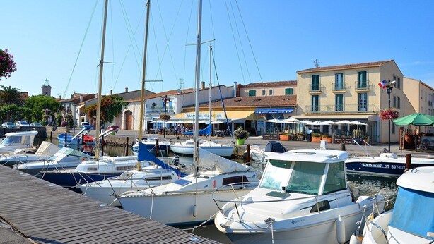 A marina with several boats docked, a wooden walkway, and buildings with shops and outdoor seating.