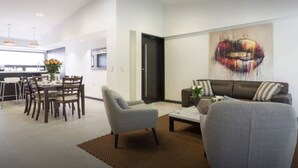 A modern living room with a sofa, armchairs, and a coffee table. There is a dining area with a table and chairs, and a kitchen in the background.
