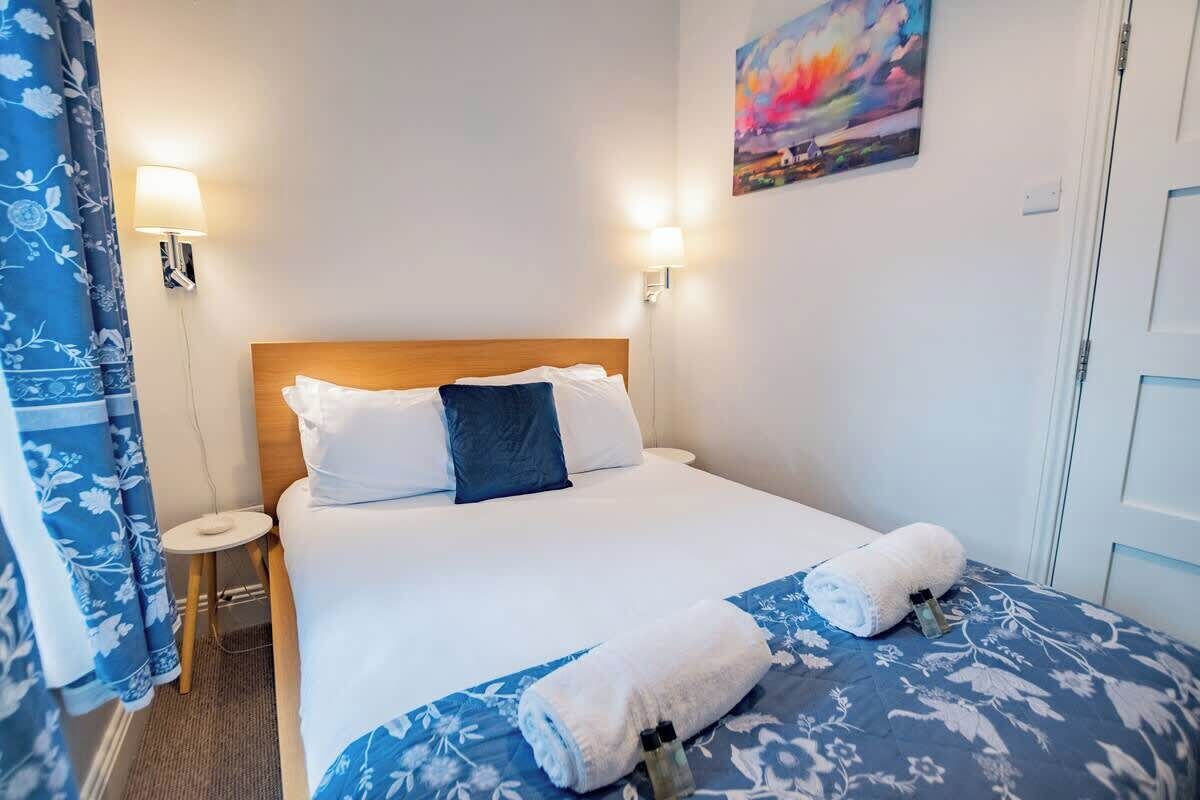 Ein Hotelzimmer mit einem Bett, zwei Nachttischlampen, einem kleinen Tisch, einem Gemälde an der Wand und einer Tür.