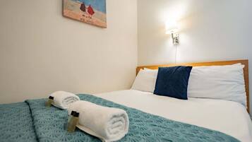 Habitación de hotel con cama, dos toallas, una manta azul, un cuadro en la pared y una lámpara fijada a la pared.