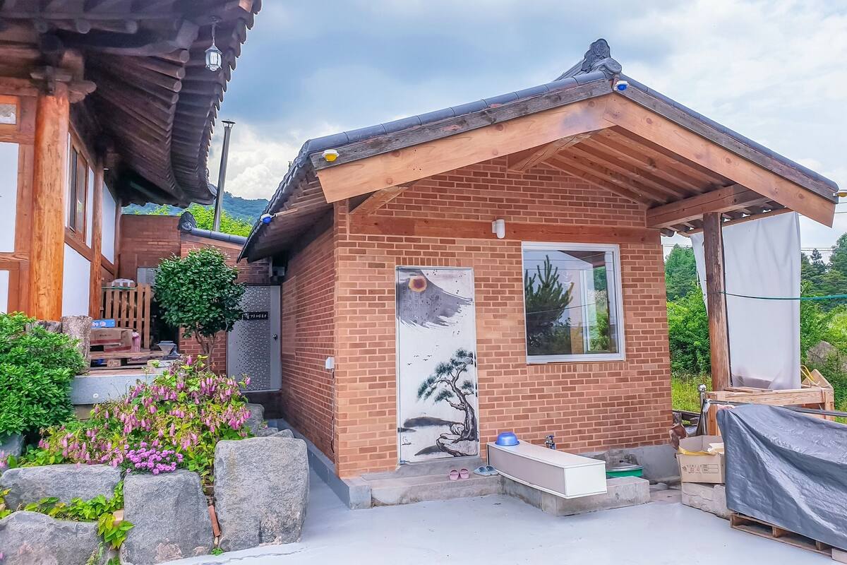 Une maison traditionnelle de style coréen, avec un toit en bois, une façade en briques et un petit jardin agrémenté de rochers et de fleurs.