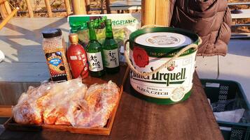 A wooden table with a large can of Pilsner Urquell beer, several bottles of beer, a jar of seasoning, a bag of vegetables, and a tray of cooked meat.
