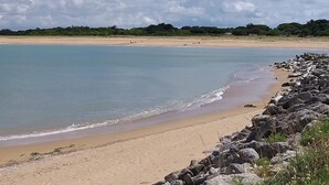 Une plage bordée d’un rivage rocheux, avec une mer calme et des arbres qui se dessinent à l’horizon sous un ciel partiellement nuageux.