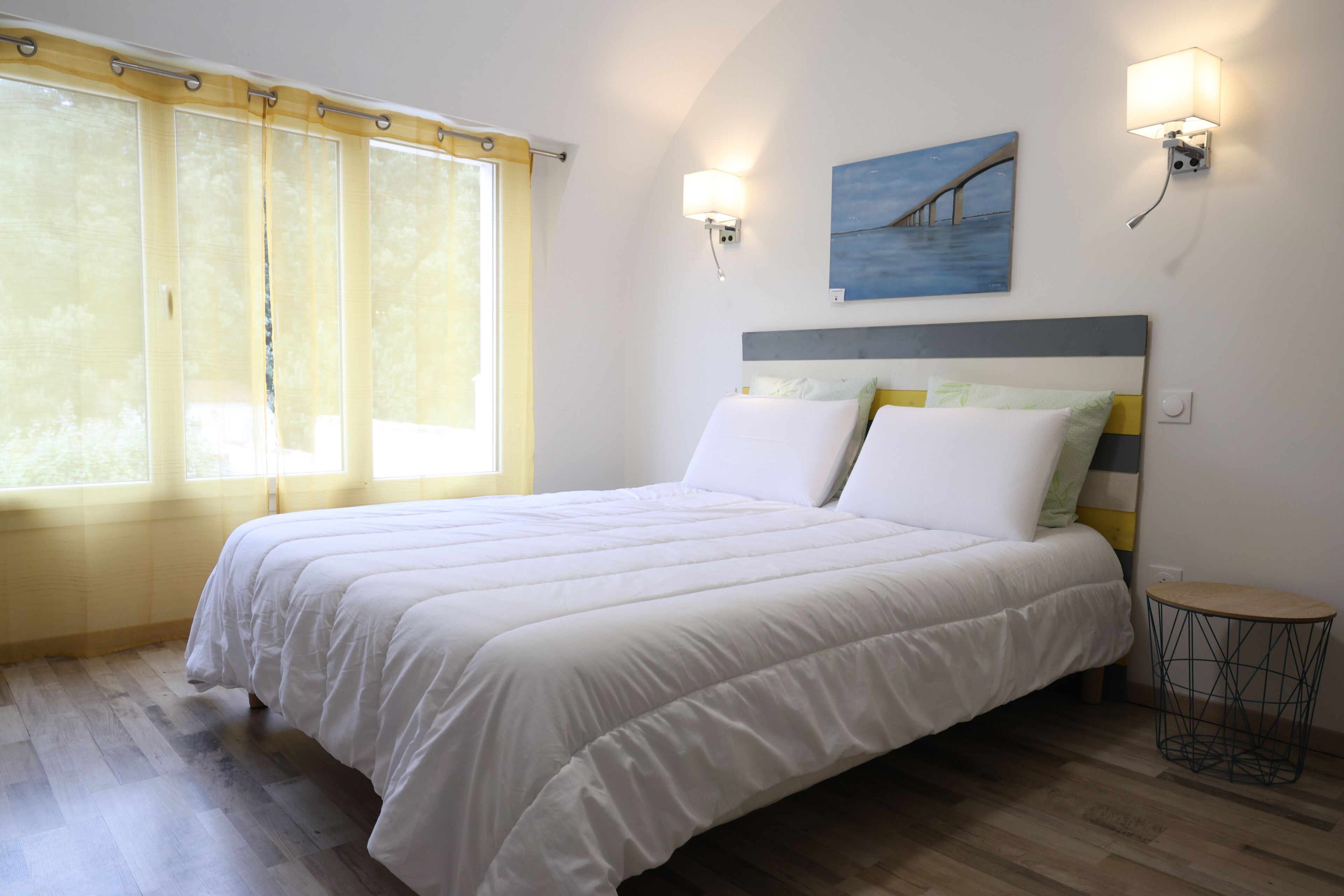 A bedroom with a bed, a window with curtains, a wall-mounted lamp, and a painting.