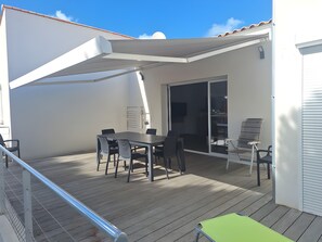 Une terrasse extérieure avec une table et des chaises, un espace salon abrité et une porte-fenêtre coulissante donnant sur un espace intérieur.