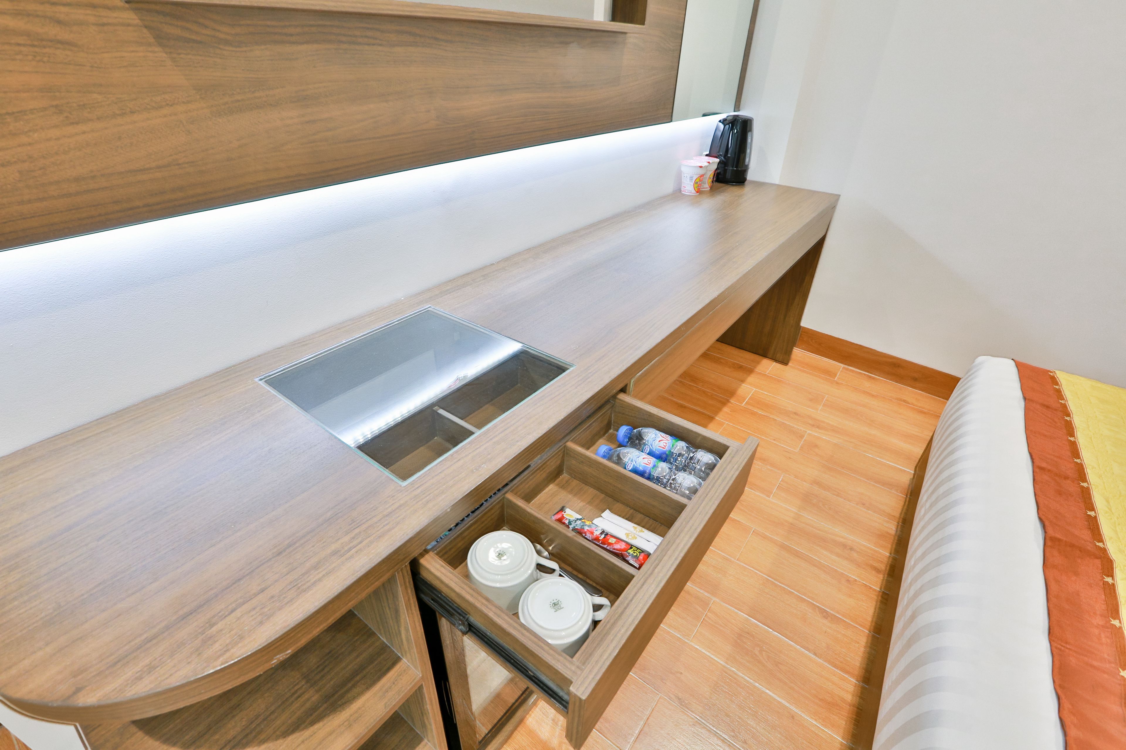 A modern hotel room with a wooden desk, a built-in sink, and a storage drawer containing kitchen items.