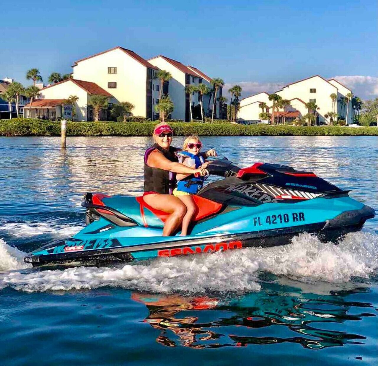 A jet ski with 'FL 4210 RR' on the side, a person and a child on it, and a body of water with houses in the background.