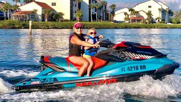 A jet ski with 'FL 4210 RR' on the side, a person and a child on it, and a body of water with houses in the background.
