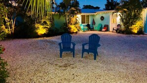 Una casa con camino de grava, dos sillas azules mirando hacia la fachada, y un porche con una silla verde y un paraguas rojo.