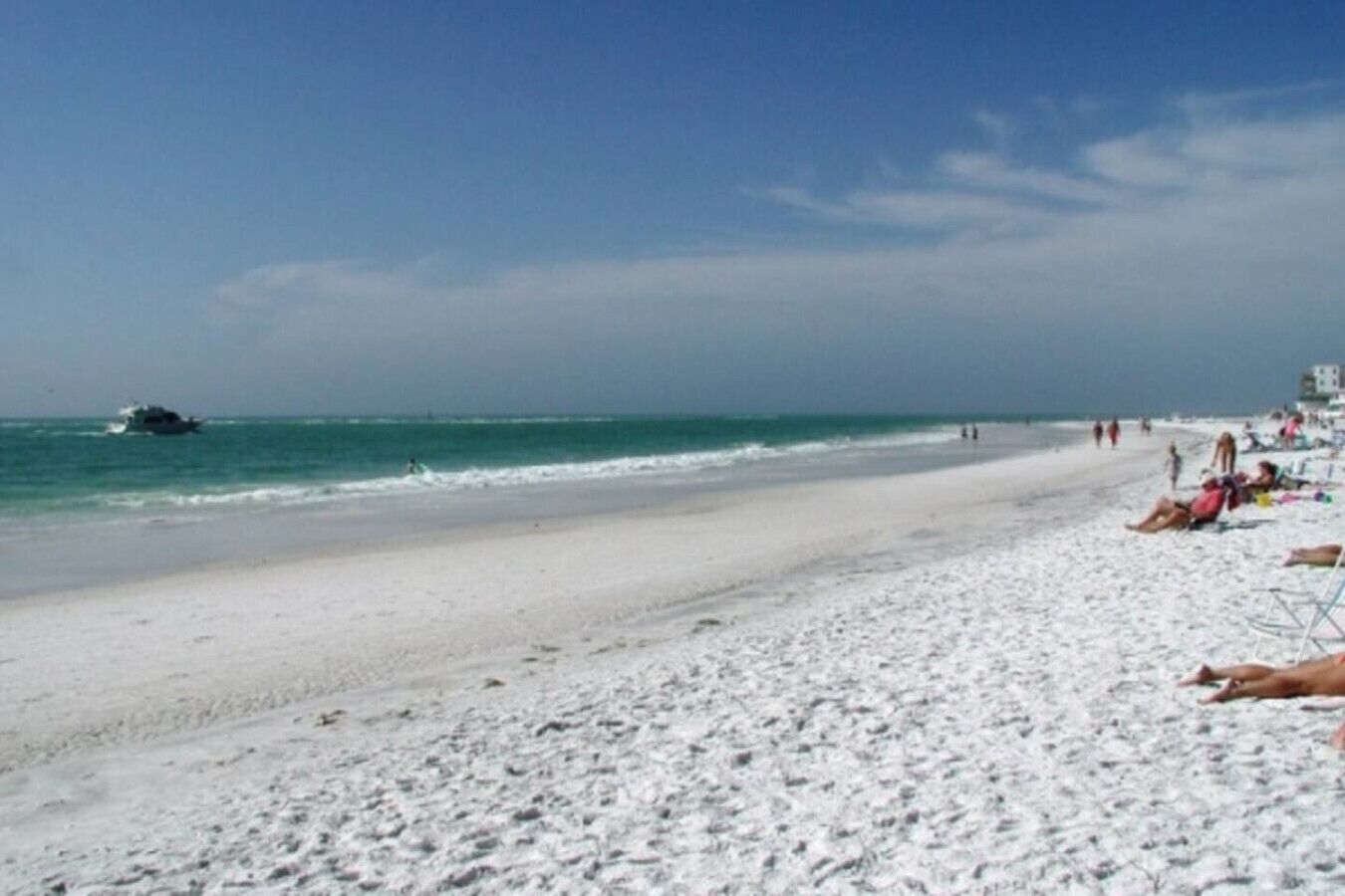Une plage avec un bateau sur l’eau, des personnes sur le sable et des bâtiments en arrière-plan.