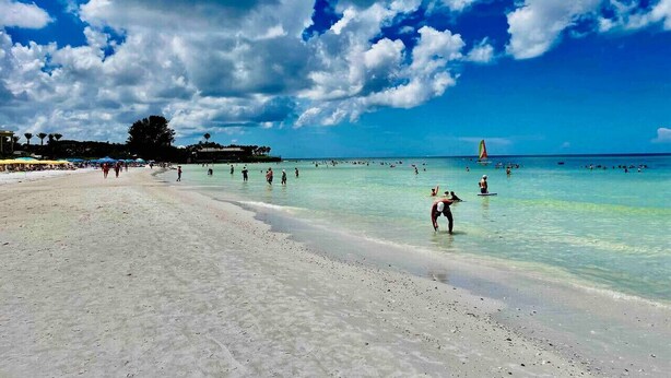 Una playa con agua turquesa cristalina, gente nadando y tomando el sol, y un velero a lo lejos.