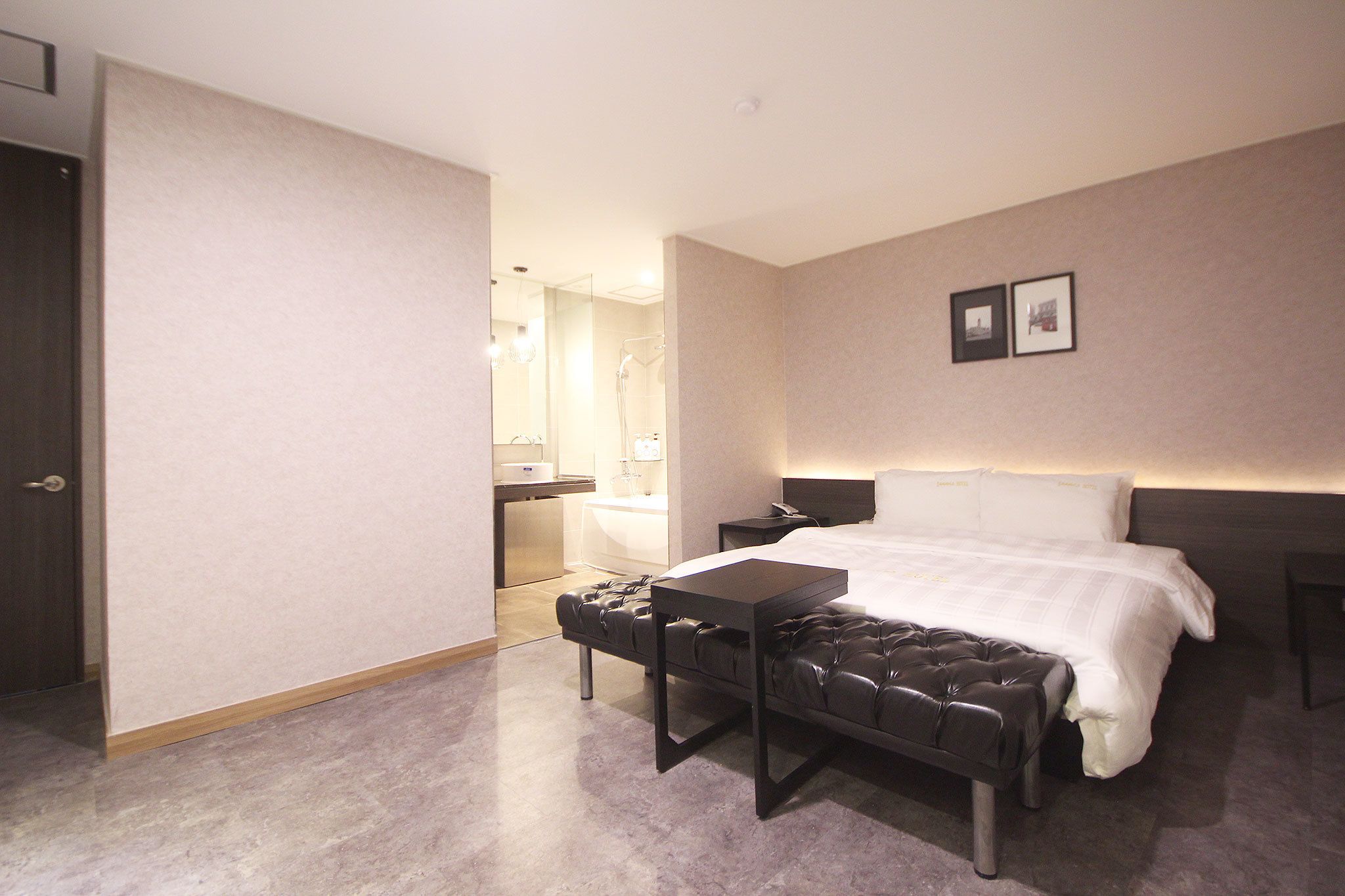 A hotel room with a bed, a bench, a bedside table, a nightstand, a bathroom with a shower, and a toilet.