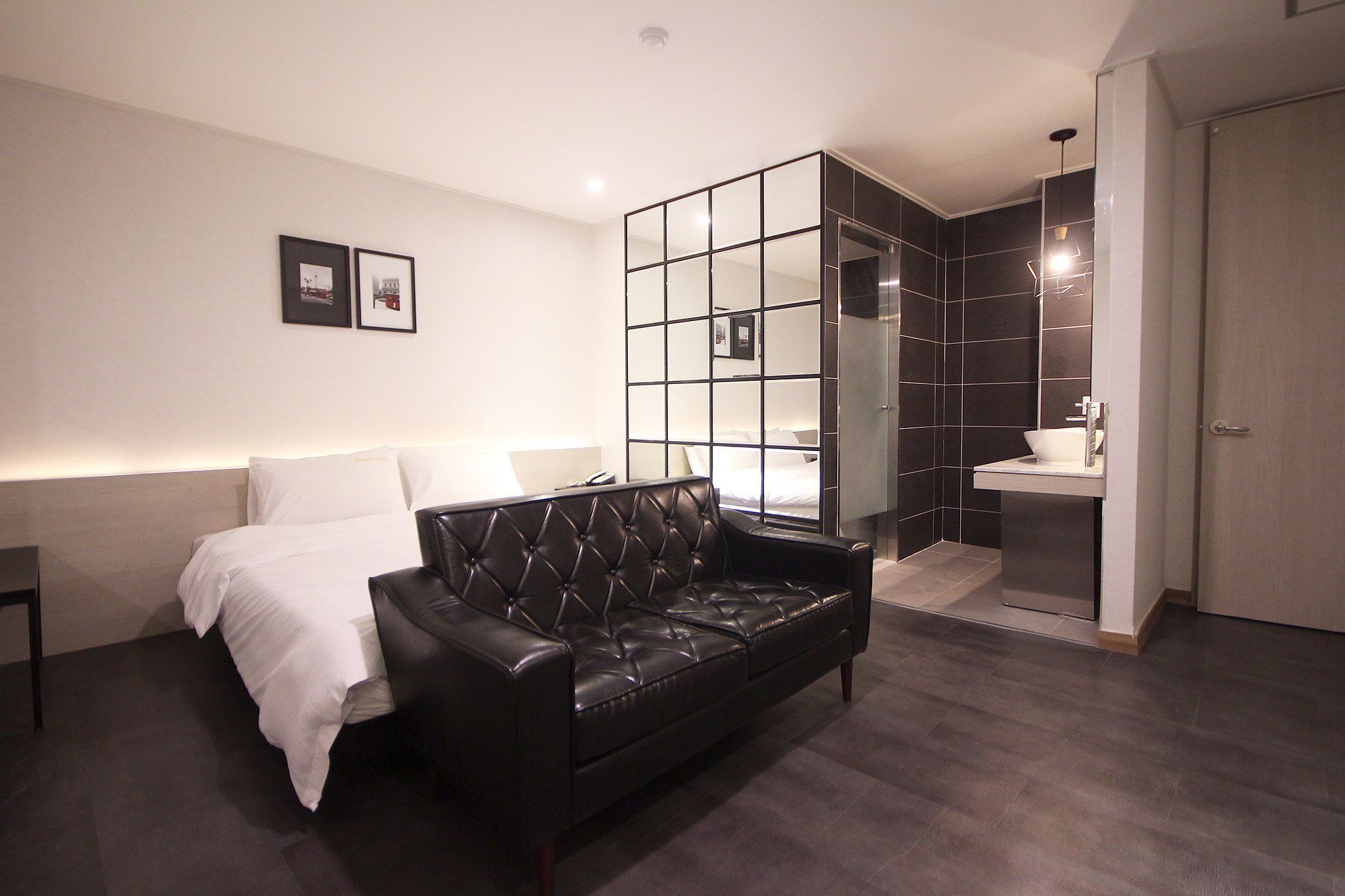 A modern hotel room with a black leather sofa, a bed with white linens, a glass partition, a bathroom with a sink, and a door.