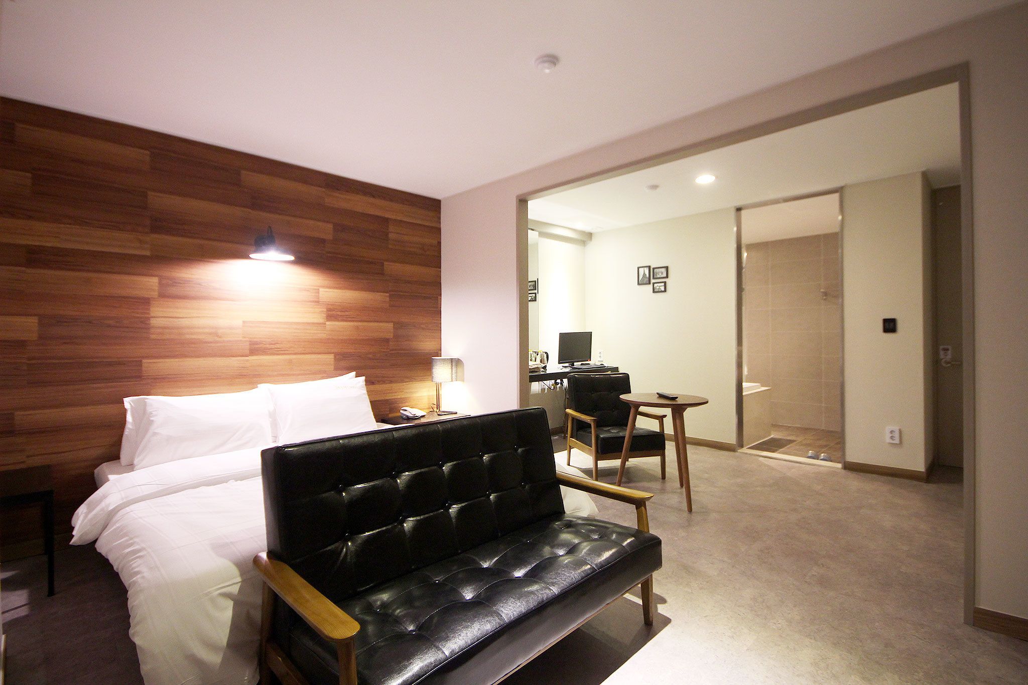 A hotel room with a wooden headboard, a black leather sofa, a bed with white linens, a desk, and a chair.
