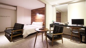 Et hotelværelse med en seng, to sorte læderstole, et rundt bord, et skrivebord med en computer og et spejl.