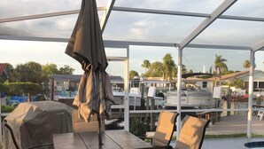 Ein Patio im Freien mit überdachtem Tisch und Stühlen, einem Grill und im Hintergrund eine Aussicht auf Boote und Palmen.