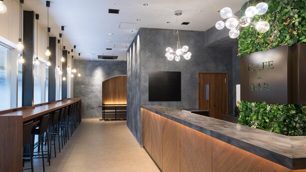 Eine moderne Bar mit einer langen Theke, Barhocker und einer grünen Wand mit dem Schild 'Cafe & Bar'.