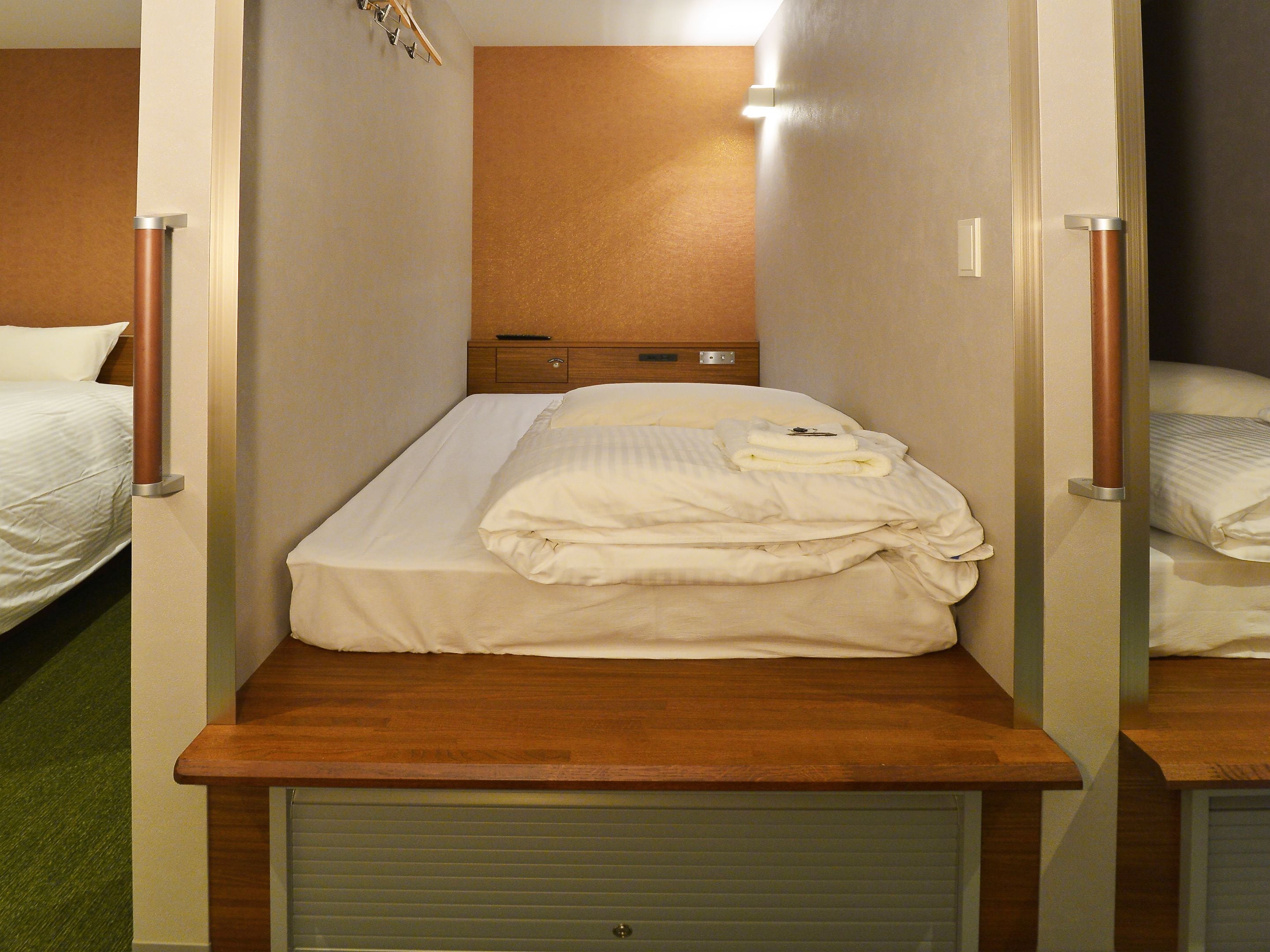 A compact hotel room with a single bed, a wooden headboard, a small bedside table, and a built-in wardrobe.