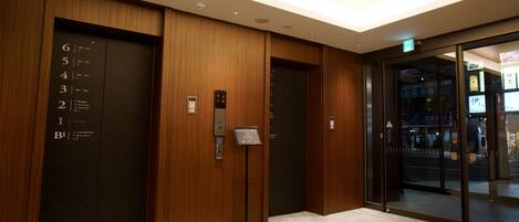 Un ascensor con los números de los pisos y las indicaciones de dirección, un cartel que señala la entrada principal y una puerta de cristal que da a la calle.