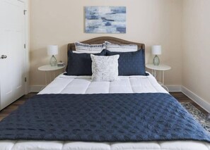 Un lit bien fait, recouvert d’une courtepointe bleu foncé, avec deux oreillers blancs et un coussin à motifs. On y trouve deux tables de chevet avec des lampes, et un tableau accroché au mur.