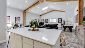 Un vaste espace de vie doté d’un haut plafond et de poutres apparentes, avec un îlot central agrémenté d’une plante, une table de billard et un coin repas comprenant des chaises et une table.
