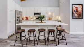 Une cuisine moderne avec des meubles blancs, un grand îlot central doté de tabourets, des appareils en acier inoxydable et une affiche de Spider-Man accrochée au mur.