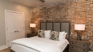 Un dormitorio con pared de ladrillo, una cama con ropa de cama blanca, dos mesitas de noche con lámparas y una puerta de madera.