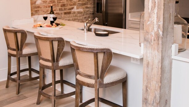Una cocina con pared de ladrillo, taburetes de madera, una isla de mármol y luces colgantes.