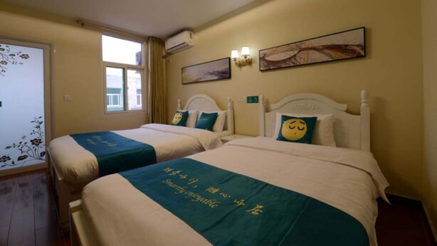 A hotel room with two beds, each with a teal bedspread and a pillow featuring a smiley face. There are wall-mounted light fixtures and a window with curtains. - Sucha Hotel Xi'an Xianyang Airport (Xianyang)