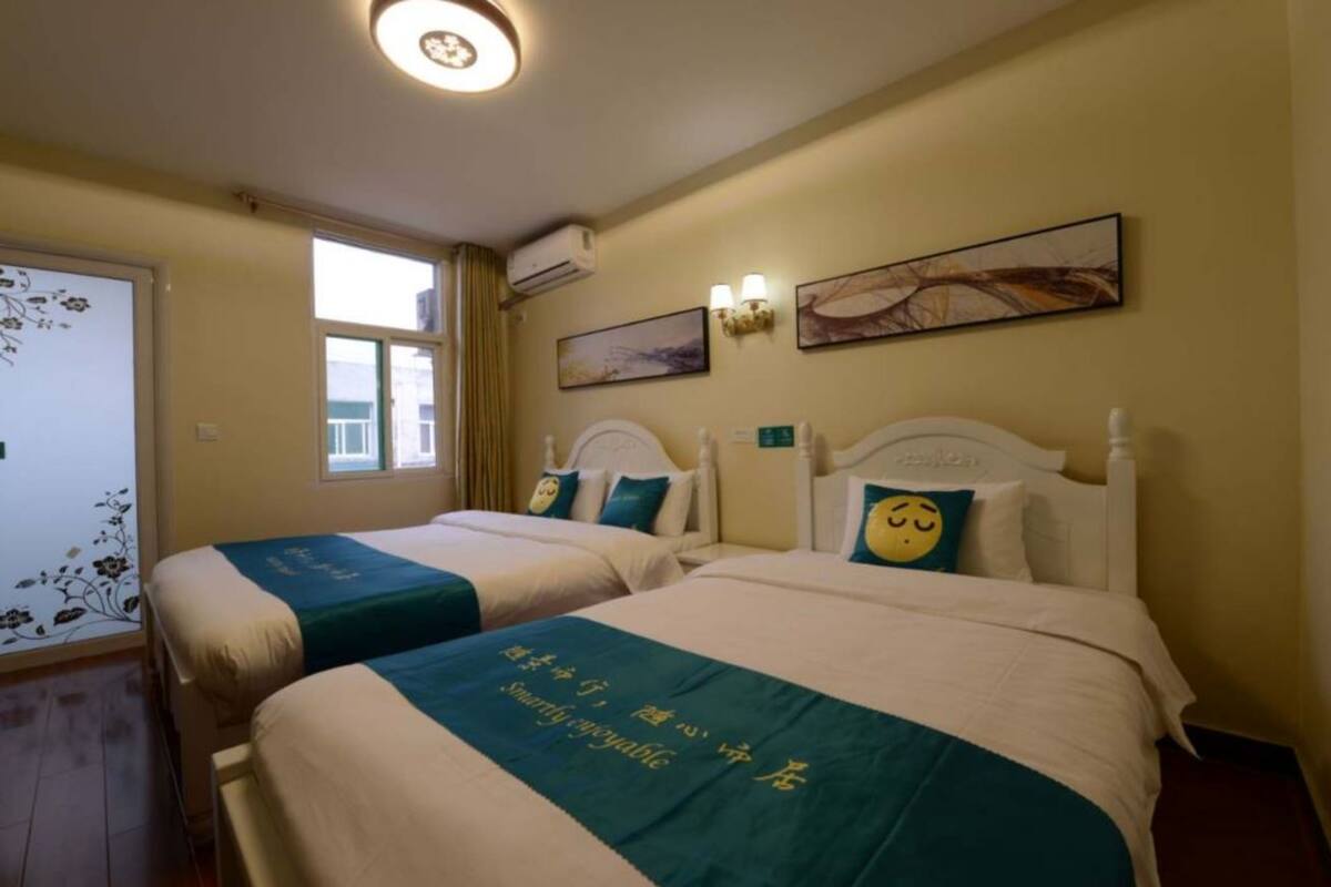 Ein Hotelzimmer mit zwei Betten, jeweils mit einer türkisblauen Bettdecke und einem Kissen mit einem Smiley. Es sind Wandleuchten und ein Fenster mit Vorhängen vorhanden.