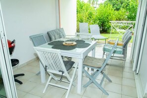 Un'area pranzo con tavolo e sedie bianche, un piccolo giardino visibile attraverso la porta in vetro e un oggetto rosso sul pavimento.