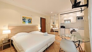 Une chambre d’hôtel avec un grand lit, une table en verre, une chaise, un bureau, un téléviseur fixé au mur et un coin cuisine visible à travers une porte coulissante.