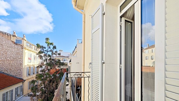 A narrow urban street with buildings on both sides, a balcony with a metal railing, and a door leading to a room with a window. - Studio Commandant André 33 by Viva Riviera (Cannes)