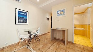 A hotel room with a dining area, a small table with chairs, a wooden side table, a framed picture of a cityscape, and a bathroom with yellow tiles. - Studio Commandant André 33 by Viva Riviera (Cannes)