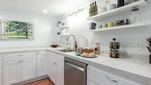 A modern kitchen with white cabinets, a stainless steel dishwasher, and a countertop with various kitchen items and a window with a view of trees. - Cozy Laurelhurst 1 Bedroom Apt Near Children’s Hospital, UW, Husky Stadium, UWMC (Seattle)