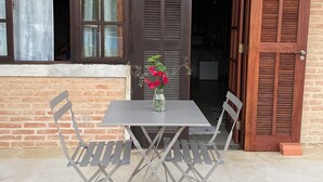 Una mesa con sillas metálicas y un florero frente a un edificio con contraventanas de madera y un muro de ladrillo.