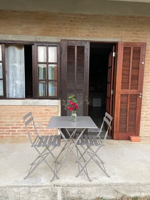 A table with metal chairs and a vase of flowers in front of a building with open wooden shutters and a brick wall. - Suites in Buzios with master barbecue (Armação dos Búzios)