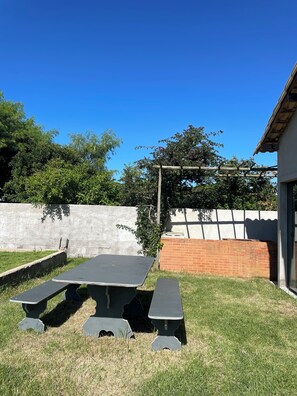 A picnic table set in a grassy yard with a white wall and a brick structure in the background. - Suites in Buzios with master barbecue (Armação dos Búzios)