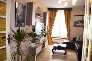 A modern living room with a wooden coffee table, a black sofa, and a TV on a wooden stand. There are potted plants and a painting on the wall.