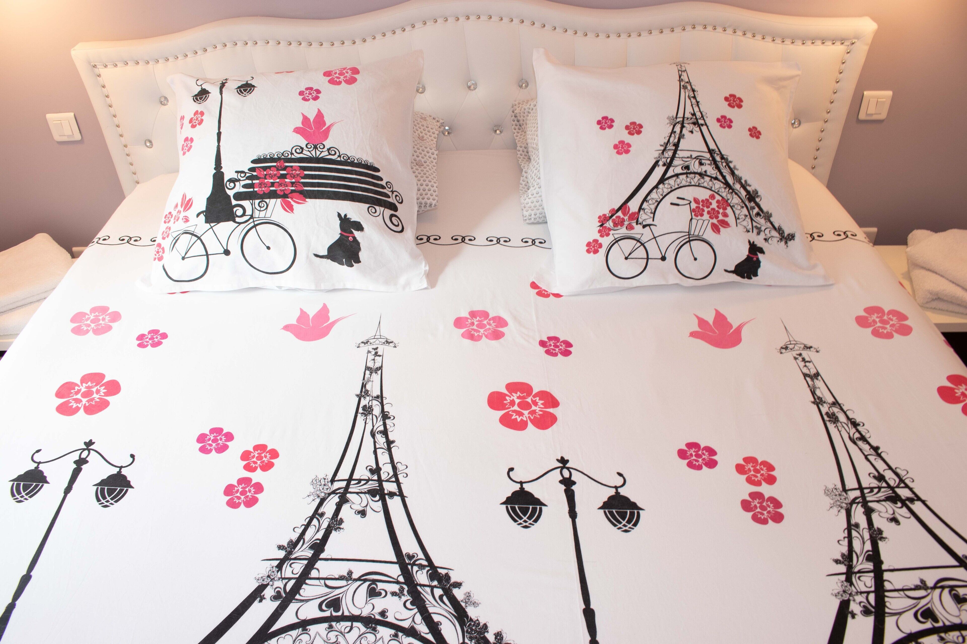 Un lit avec une literie blanche et des coussins décoratifs représentant la Tour Eiffel et des fleurs.