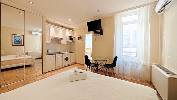 A hotel room with a bed, a small kitchenette, a dining area with a table and chairs, a TV mounted on the wall, and a window with curtains. - Studio Commandant André 13 by Viva Riviera (Cannes)