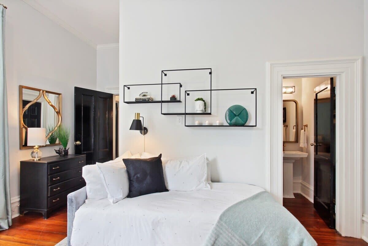 Quarto com cama, cômoda, espelho e prateleiras fixas com objetos decorativos.