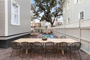 A backyard with a wooden table and chairs, a swimming pool, and a large tree.