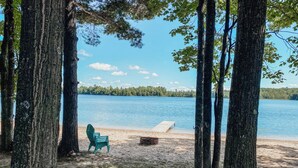 Un cadre paisible au bord d’un lac, avec une jetée en bois qui s’avance sur l’eau, entouré de grands arbres et agrémenté d’une chaise verte sur la plage de sable.