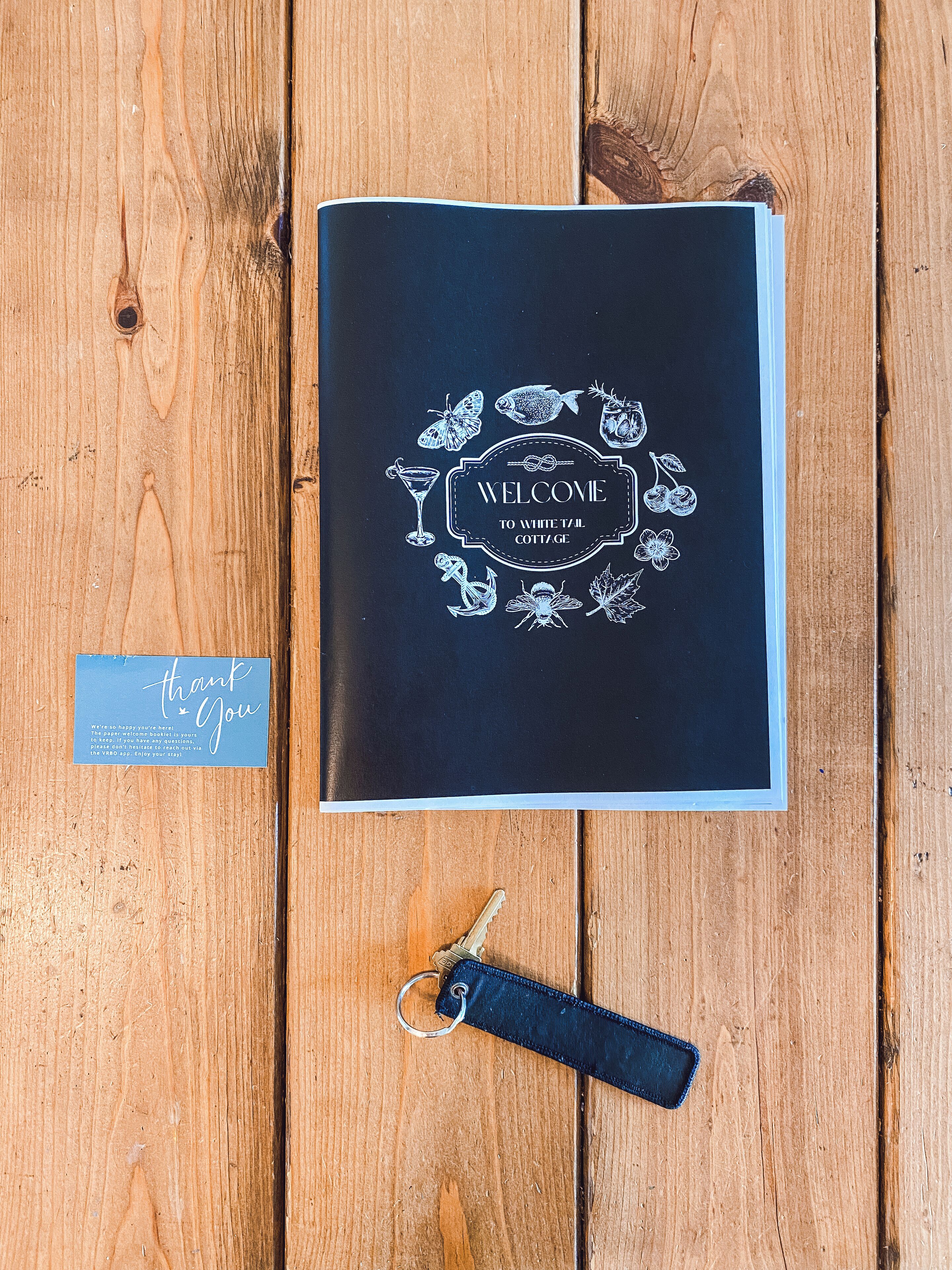 Un petit guide d’accueil noir avec des illustrations blanches représentant des aliments et des boissons, un porte-clés et une carte de remerciement, le tout posé sur une table en bois.