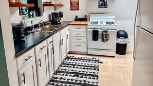 Une cuisine de taille réduite avec des meubles blancs, une cuisinière, un évier et un réfrigérateur. On trouve des étagères en bois au-dessus du plan de travail et une poubelle noire sur le côté.